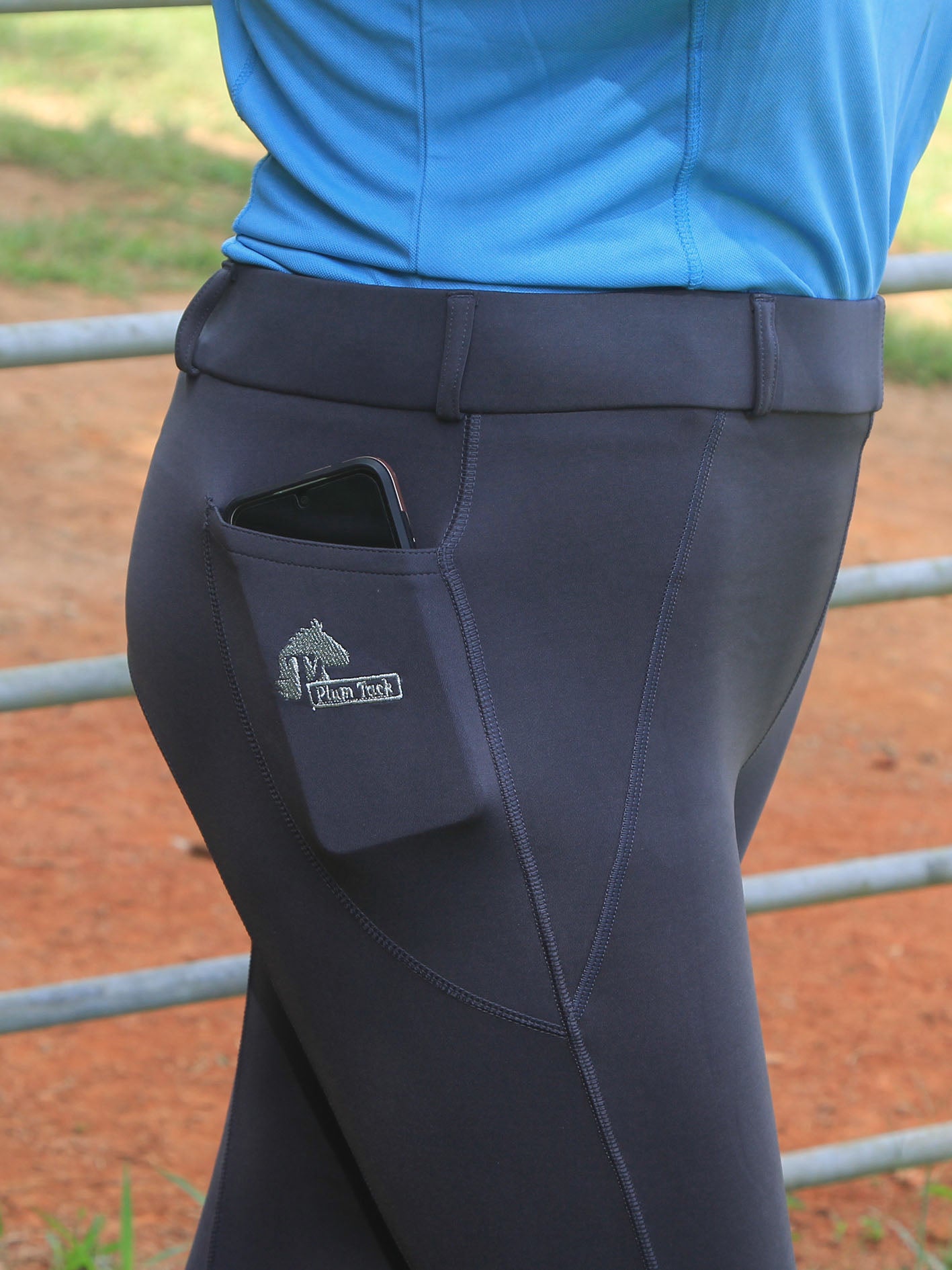 Dark grey riding pants with a phone pocket featuring a logo, worn by a person in a blue shirt. showing phone in pocket